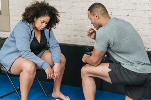 Sportive black personal coach demonstrating exercise to exhausted plump African American female in activewear sitting on chair during workout in gym