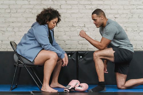 Side view of tired African American plump female sitting near muscular personal couch in gym with gloves during boxing workout