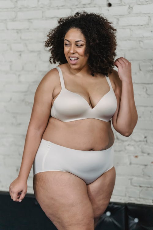 Positive plus size African American female with curly hair wearing white bra and panties standing near brick wall in studio