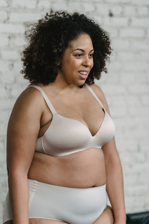 Cheerful plus size African American female with curly hair wearing bra and panties standing against white brick wall in room
