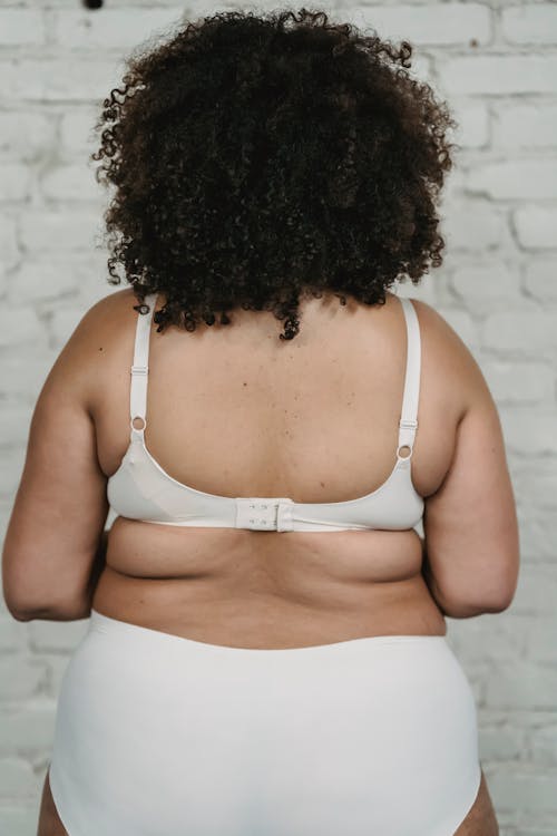 Back view of anonymous plus size female with curly hair wearing underpants and bra standing near white brick wall in studio