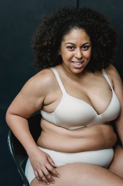 Cheerful overweight African American female in lingerie smiling and looking at camera while sitting on chair against black background in studio