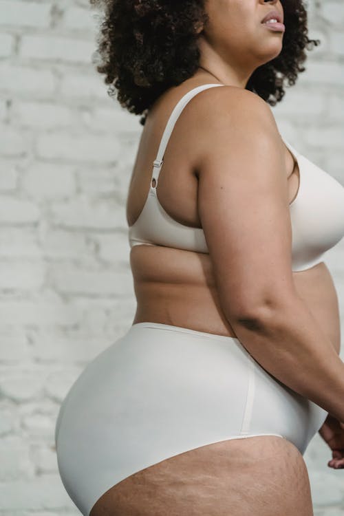 Side view of plus size African American female with curly hair wearing underwear standing near white brick wall in room