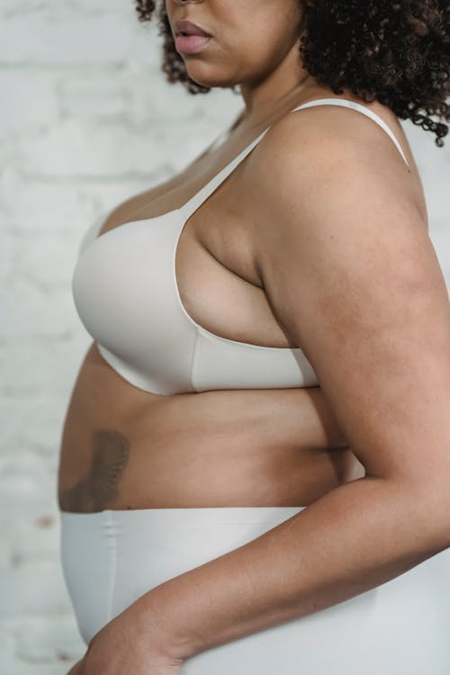 Side view of calm African American overweight female with dark hair and tattoo wearing white underwear standing near white brick wall