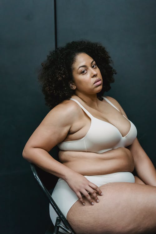 Serious adult plump ethnic female in white underwear looking at camera while sitting on chair in studio near gray wall