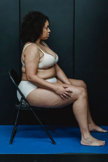 Side view of confident adult ethnic plump woman in white underwear sitting on chair on blue floor in light studio near gray wall