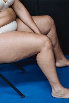 Crop faceless plump female in white underwear sitting on chair on blue floor in light studio on black background