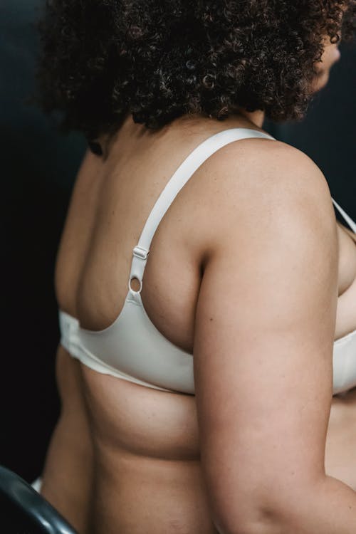 Crop faceless plump woman in white underwear sitting on chair on black background in light studio