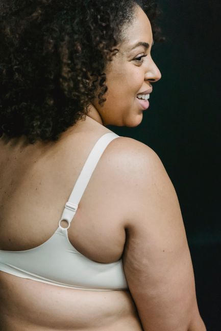 Smiling adult plump ethnic female in white bra on dark background in bright studio