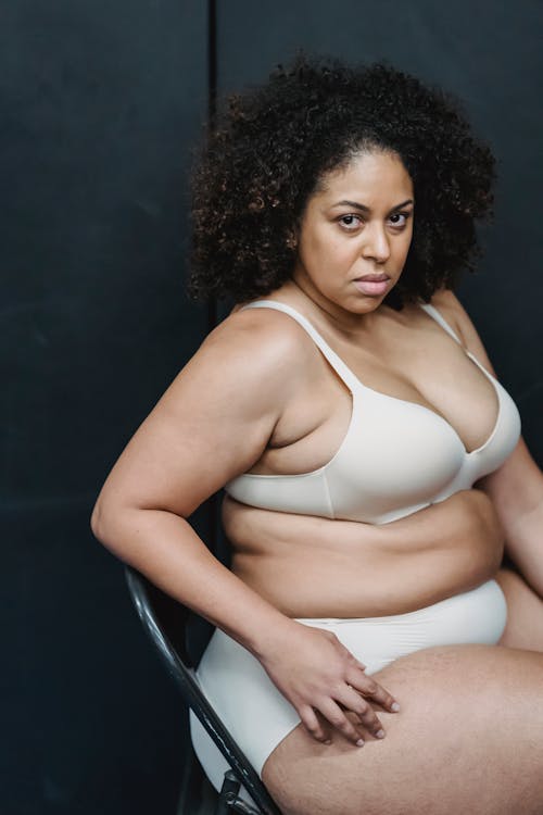 Serious adult plump ethnic female in white underwear looking at camera while sitting on chair near gray wall in bright studio