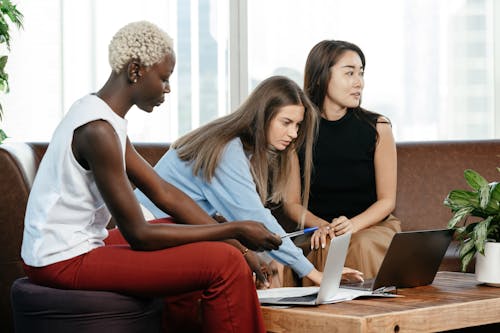 Side view of multiracial female coworkers typing on netbooks while sitting at table on couch during teamwork in modern office