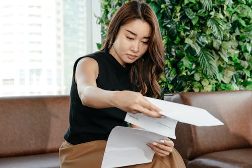 Focused Asian female turning pages of document while sitting on sofa during paperwork in modern workspace with green deciduous plant
