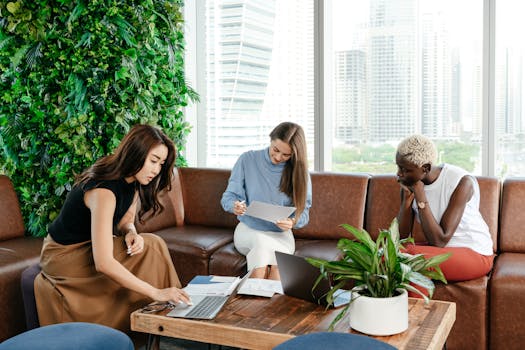 Diverse female coworkers working with documents and browsing modern netbooks while sitting on sofa near wooden desk in modern workspace