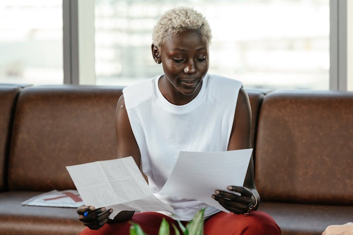 Concentrated African American female comparing important papers of report on leather sofa in office