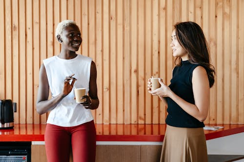 Free Happy multiracial women with cups of coffee smiling and discussing job in contemporary office Stock Photo