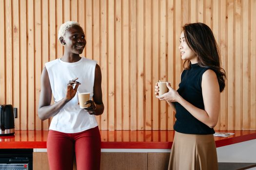 Happy multiracial women with cups of coffee smiling and discussing job in contemporary office