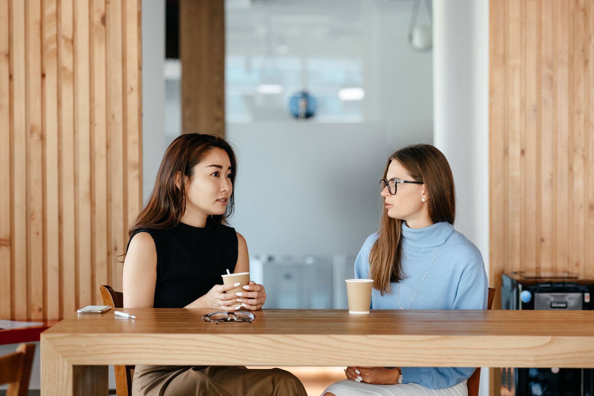 Two women enjoy a coffee and chat at a modern indoor cafe setting.