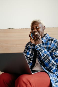 Relaxed young woman in checkered shirt chatting on phone while using laptop indoors.