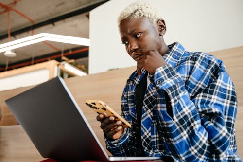 Young serious African American female in blue checkered shirt looking at screen of netbook while texting message on mobile phone