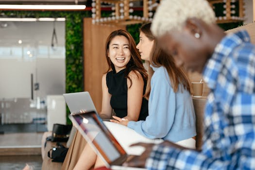 A group of diverse professionals engaging in conversation and work on laptops in a modern office setting.