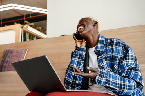 Free From below of positive African American female in fashionable bright blue shirt speaking on mobile phone while working with netbook Stock Photo