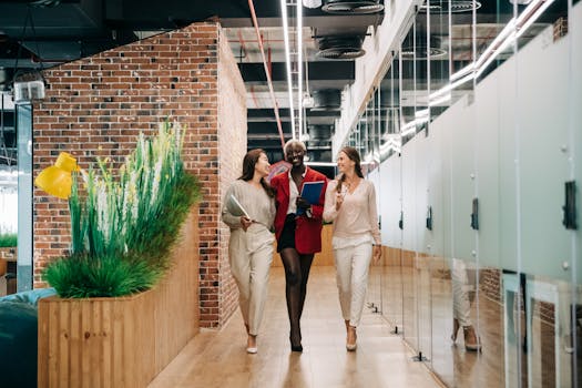 Three diverse businesswomen walking and smiling in a bright modern office space, engaging in positive conversation.