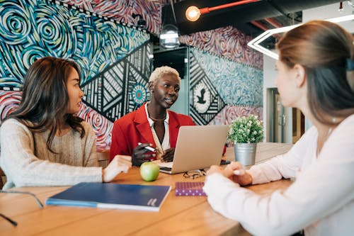 Pensive multiracial coworkers in stylish clothes sitting at table with laptop and notebooks and discussing details of project in modern workspace