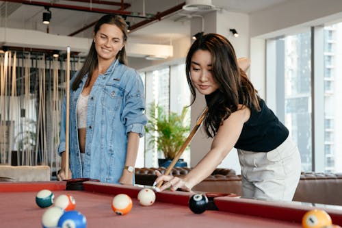 Free Positive diverse women in casual clothes playing billiard together in spacious room in club in daytime Stock Photo