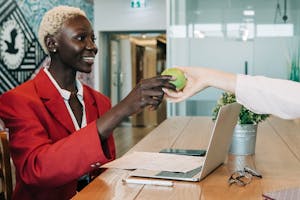 Side view of cheerful African American businesswoman in stylish clothes sitting at wooden table with netbook and documents and taking green apple from coworker in modern workspace