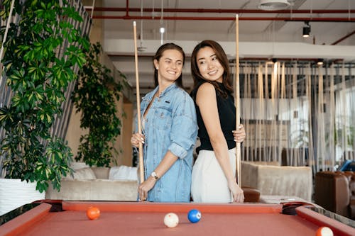 Free Cheerful multiethnic friends in casual clothes standing with billiard cues while playing pool game in spacious room in daytime Stock Photo
