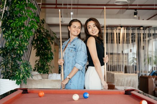 Cheerful multiethnic friends in casual clothes standing with billiard cues while playing pool game in spacious room in daytime