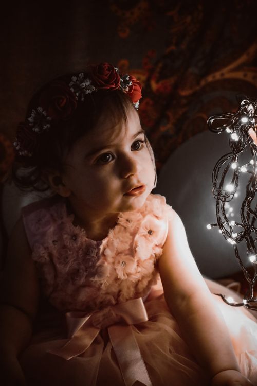 Free A young girl wearing a floral dress and flower crown gazing at glowing fairy lights indoors. Stock Photo