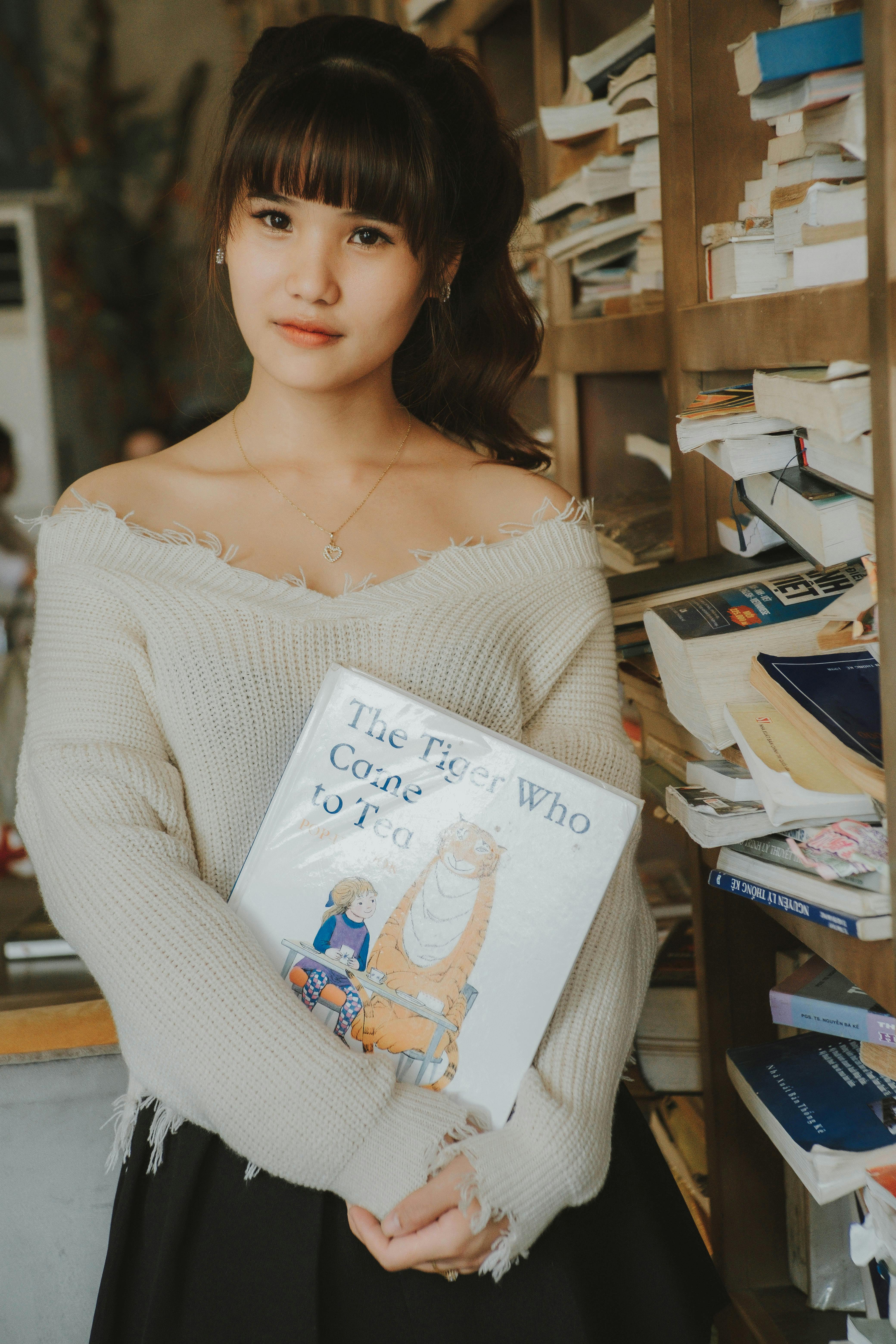 Asian woman with bangs holding 'The Tiger Who Came to Tea' indoors, surrounded by books.