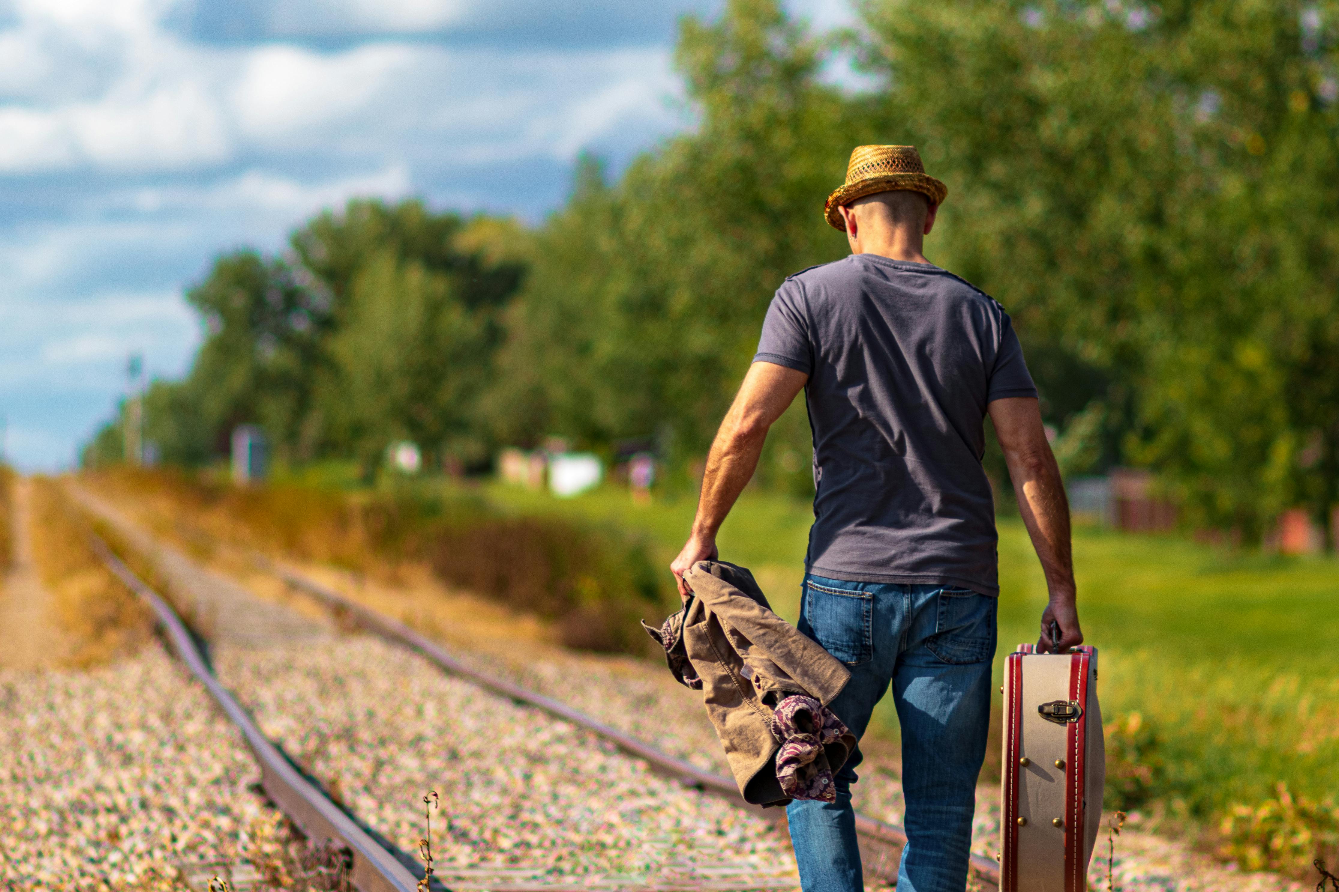 Man walking down railroad tracks holding a suitcase and coat on a sunny day.