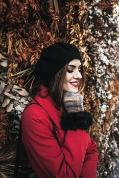 A woman wearing a red coat and black beret smiles in front of a textured autumn wall.