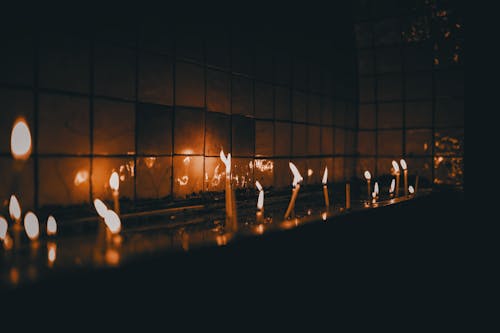 Little burning thin fragile candles near glass wall reflecting flame in dark room