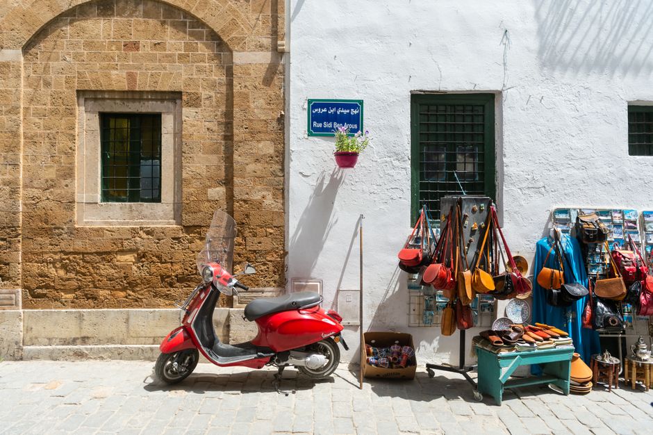 Modern red scooter parked near aged residential building on sidewalk near local market with assorted handbags in old sunny town