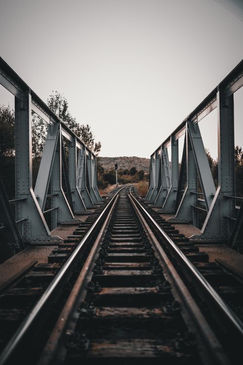 Free Straight railroad tracks amidst metal construction of bridge going through mountainous terrain against cloudless sunset sky Stock Photo