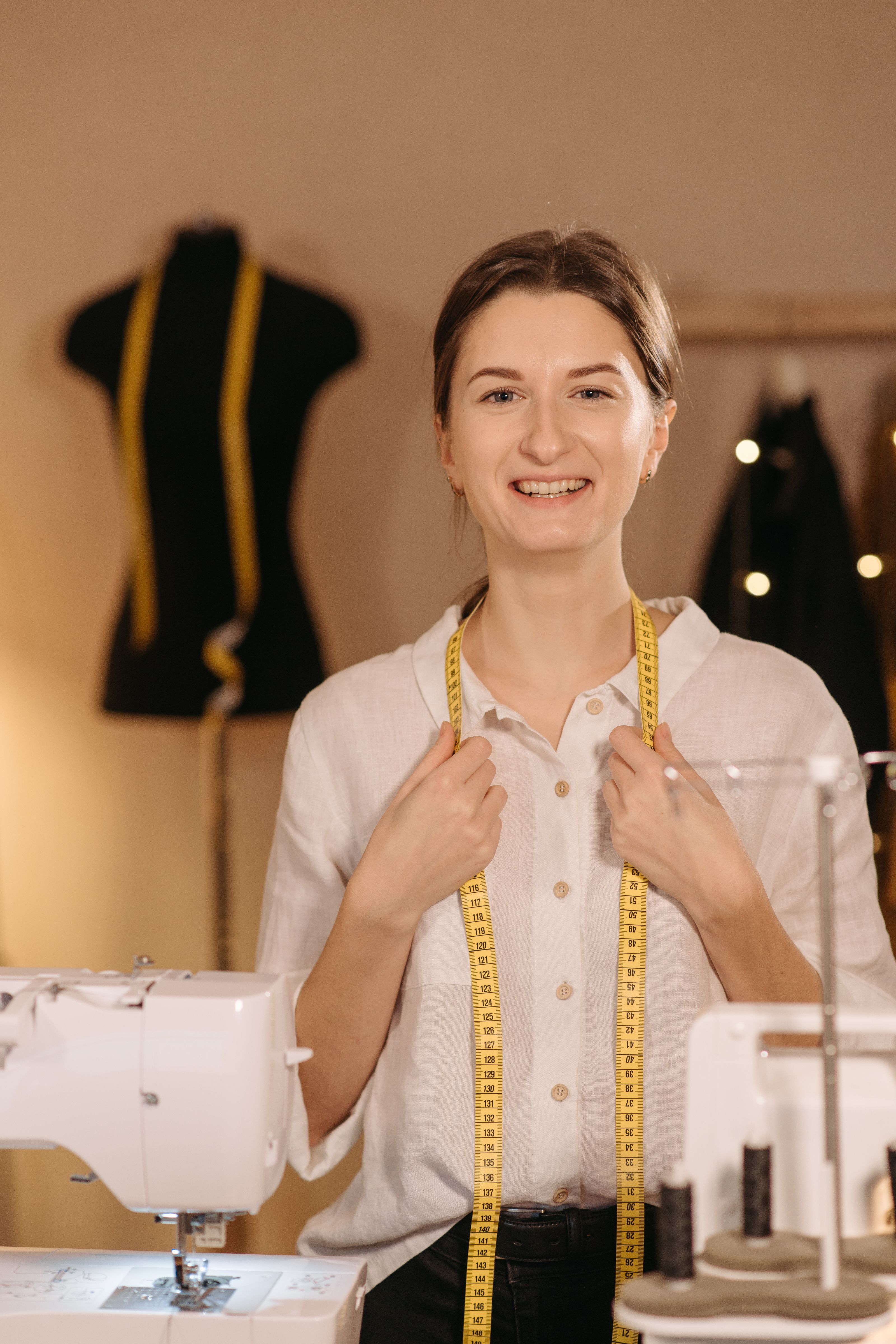 Uma feliz costureira em um estúdio de costura com uma fita métrica ao redor do pescoço e máquinas de costura à vista.