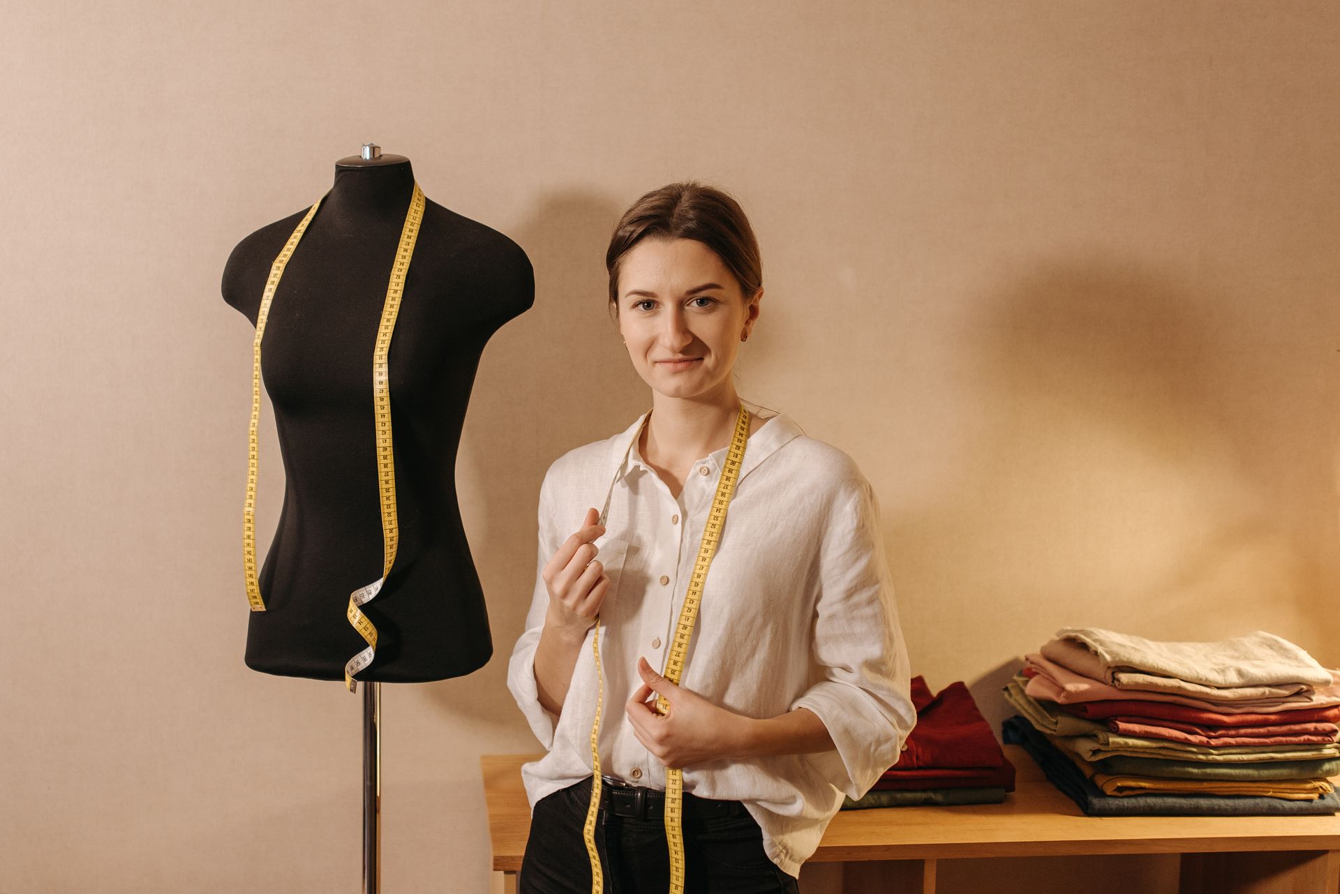Dressmaker Smiling in White Long Sleeve Blouse Standing Beside Black Manikin with Measuring Tape Hanging on Her Neck