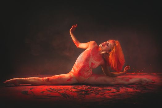 Contemporary dancer in a bodysuit performs a split surrounded by vibrant red powder and smoke.