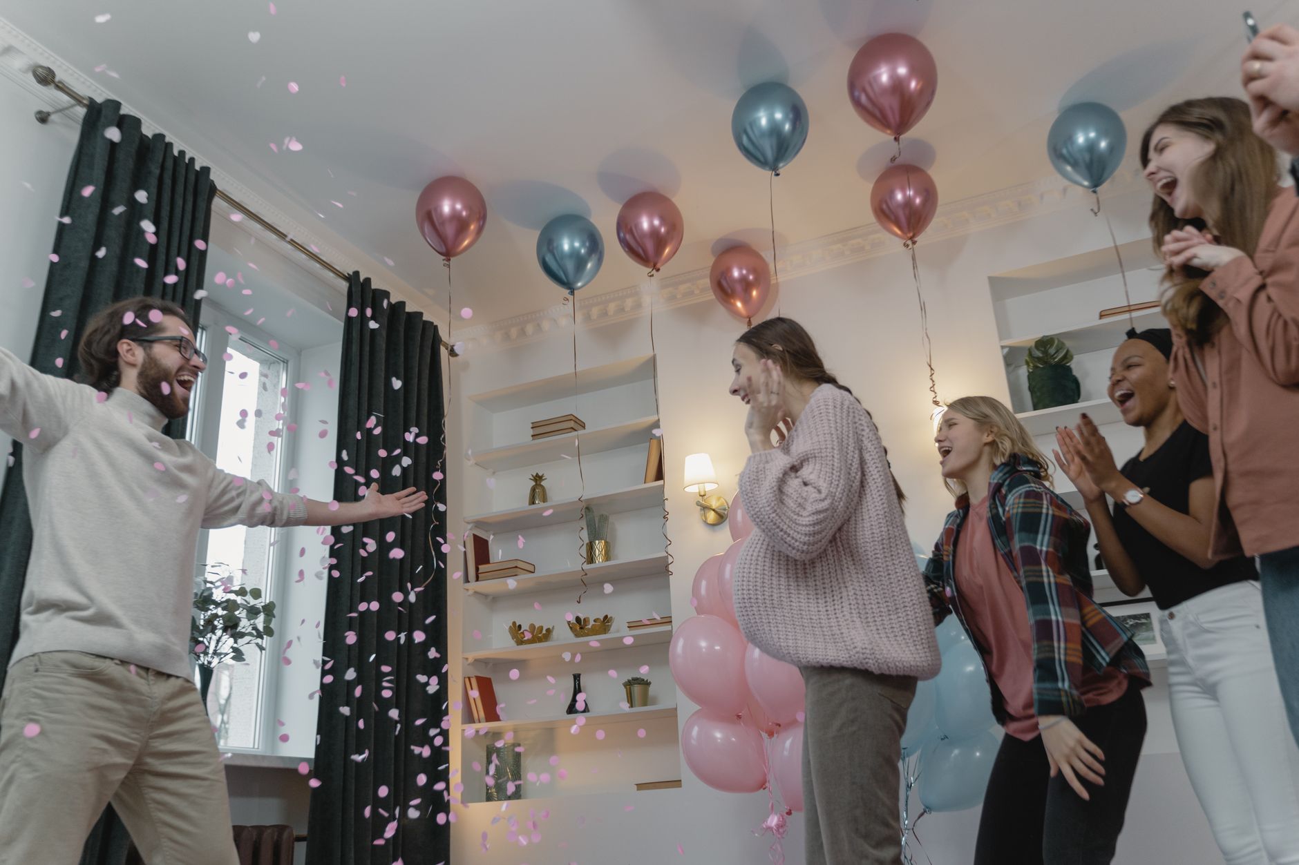 Group of friends celebrating a joyful baby shower with balloons and confetti indoors.