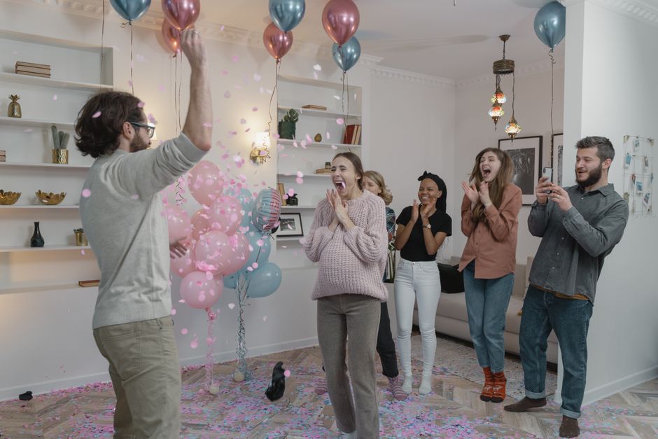 A group of excited people celebrating a gender reveal with balloons and confetti indoors.