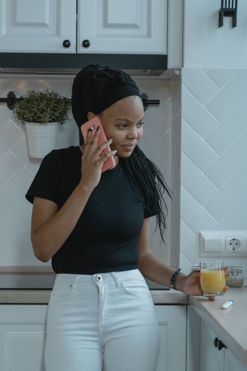 Free Young woman in kitchen holding a phone and glass of juice, looking thoughtful. Stock Photo