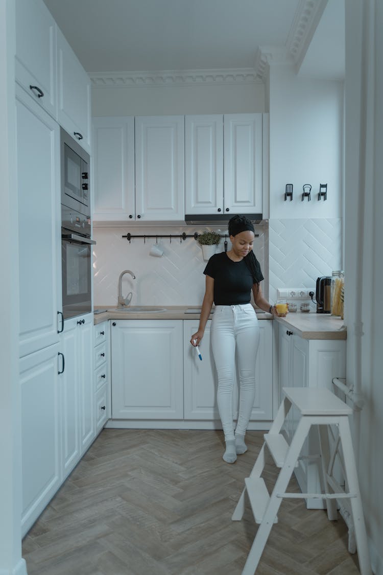 Woman In Black Tank Top And White Pants Standing In Front Of White Wooden Kitchen Cabinet