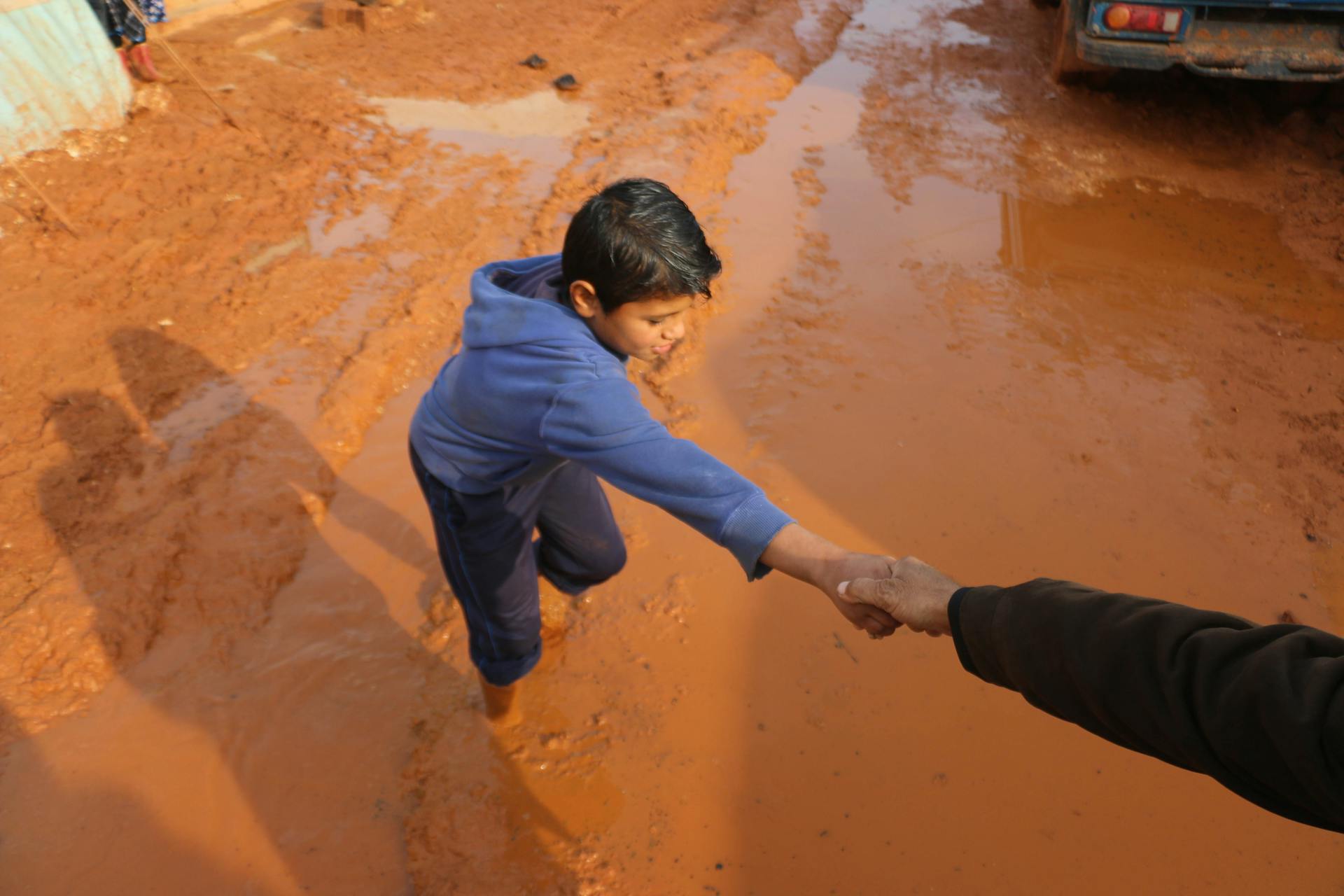 High angle of crop person holding hands with ethnic boy stuck in dirty puddle in poor village