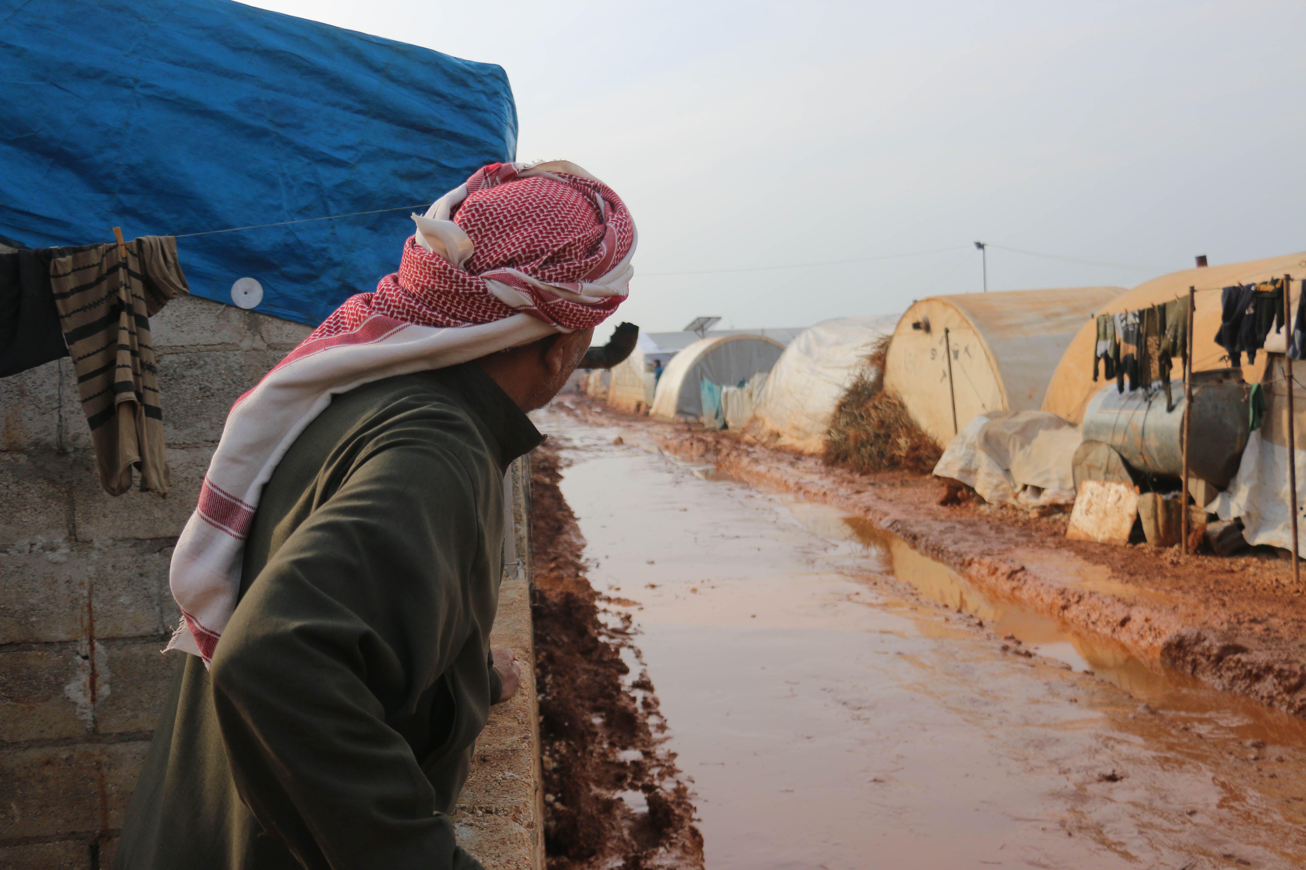 伝統的な服装をした男性が、シリアのイドリブ難民キャンプの泥だらけの道を見つめています。