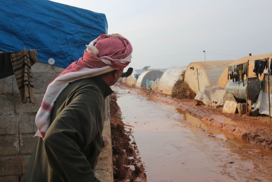 伝統的な服装をした男性が、シリアのイドリブ難民キャンプの泥だらけの道を見つめています。