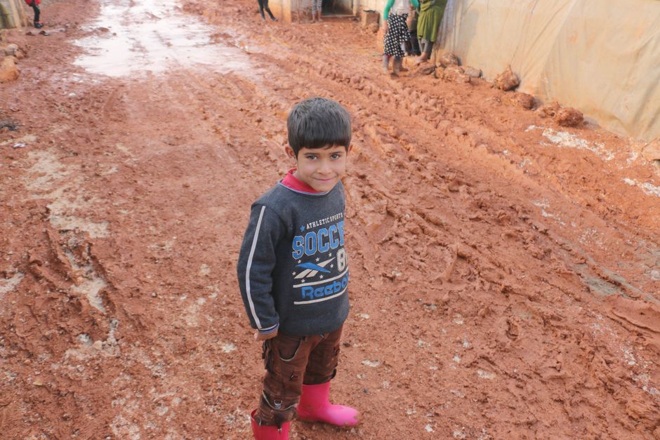 From above of glad ethnic kid inc casual wear standing on dirty path near tents in poor district of refugee camp