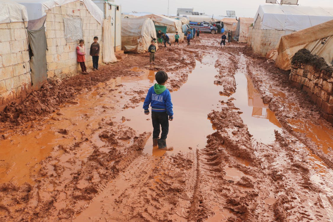 Free Group of children standing on dirty wet ground with puddles between old tents in refugee camp with in poor settlement Stock Photo